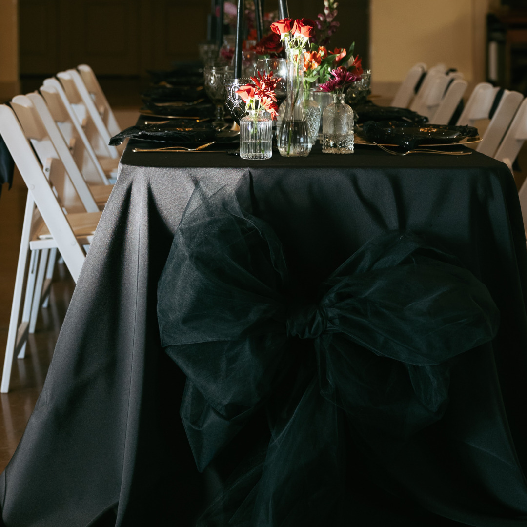 A table with a black cloth, a black bow on the front, and a table set-up on top.